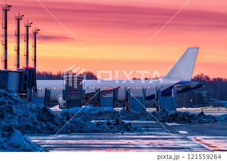 A passenger plane stands on the airport near a row of aircraft mobile gangway against the backdrop of a picturesque crimson sunset A passenger plane stands on the airport near a row of aircraft mobile gangway against the backdrop of a picturesque crimson sunset 121559264