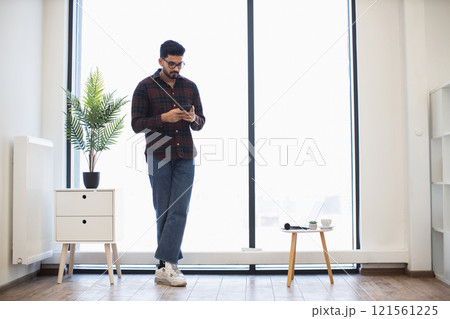 Young Indian man in casual attire using smartphone in modern office environment near large window with natural light. 121561225