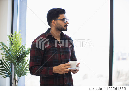 Indian man in his 30s wearing glasses holds coffee cup by window. He appears thoughtful and relaxed in casual environment with indoor plant adding natural touch. 121561226