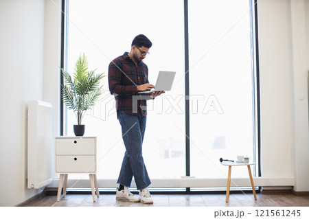 Young Indian male standing in bright office using laptop, showcasing modern work environment. Features include plant decor, wooden furniture, large window. 121561245