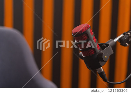 Red metal mesh professional audio microphone with black body is mounted on pantograph with connected wire against silhouette of gray chair and blurred background with orange black alternating stripes 121563305