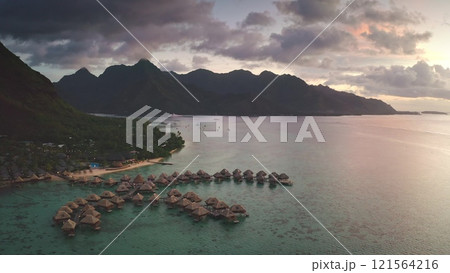 Aerial view of luxury overwater bungalows in turquoise lagoon at sunset reflecting clouds and dramatic sky with lush mountains in background in Moorea, French Polynesia. Remote wild nature paradise Aerial view of luxury overwater bungalows in turquoise lagoon at sunset reflecting clouds and dramatic sky with lush mountains in background in Moorea, French Polynesia. Remote wild nature paradise 121564216