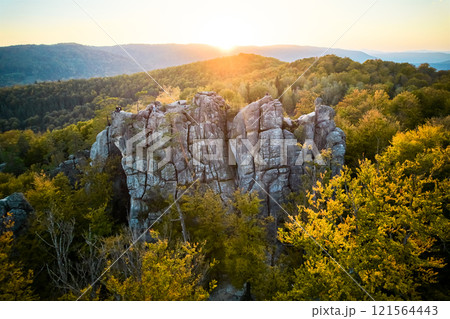 Aerial view of sun sets behind rugged rock formation, casting warm glow over landscape. Trees in autumn colors surround rocks, creating vivid contrast. Dovbush Rocks, Carpathian mountains, Ukraine. Aerial view of sun sets behind rugged rock formation, casting warm glow over landscape. Trees in autumn colors surround rocks, creating vivid contrast. Dovbush Rocks, Carpathian mountains, Ukraine. 121564443