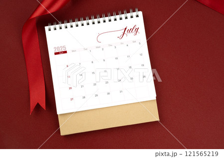 July 2025 desk calendar and red ribbon on red background. 121565219