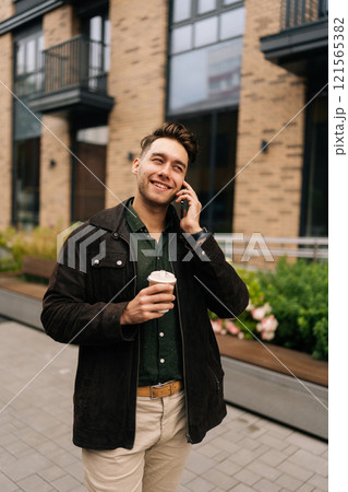 Vertical portrait of cheerful man drinking tasty coffee and talking on smartphone while walking in city street by modern urban buildings, enjoying pleasant conversation during break outdoors alone. 121565382