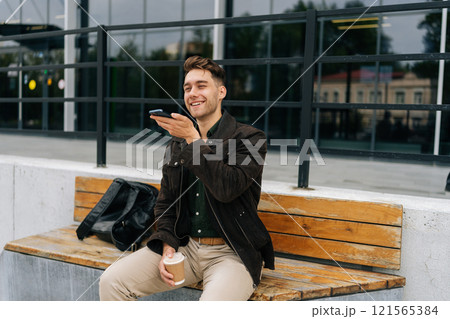 Successful businessman sitting on bench outdoors, holding takeaway coffee and recording voice message on smartphone, enjoying break time near office building. Concept of successful city lifestyle. Successful businessman sitting on bench outdoors, holding takeaway coffee and recording voice message on smartphone, enjoying break time near office building. Concept of successful city lifestyle. 121565384