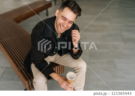 Top view of cheerful man sitting on city bench, enjoying coffee break in vibrant urban setting, laughing while savoring moment and embracing simple pleasures of life. Concept of urban lifestyle. 121565443