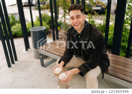 Cheerful handsome young man in casual attire enjoying peaceful moment on park bench in city, holding two coffee cups and laughing looking at camera, surrounded by green trees on beautiful summer day. 121565444