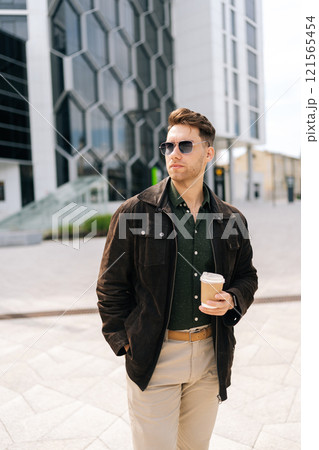 Vertical portrait of handsome bearded young man in stylish sunglasses holding takeaway coffee standing outside contemporary office building. Concept of modern successful city lifestyle. Vertical portrait of handsome bearded young man in stylish sunglasses holding takeaway coffee standing outside contemporary office building. Concept of modern successful city lifestyle. 121565454