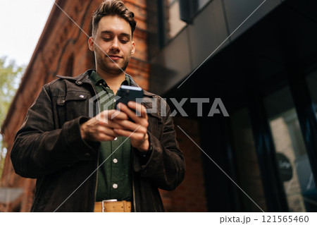 Low-angle view of handsome bearded man standing on city street, using smartphone to check messages, browse internet, stay connected enjoying urban environment. Concept of urban lifestyle. 121565460