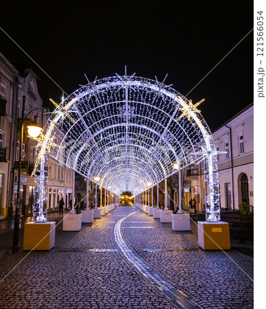 A magical winter scene of a city street adorned with twinkling Christmas lights, creating a tunnel of light. People are walking through the festive atmosphere. 121566054