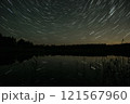 Star trails illuminate the night sky over a calm lake surrounded by a forest during a serene evening 121567960