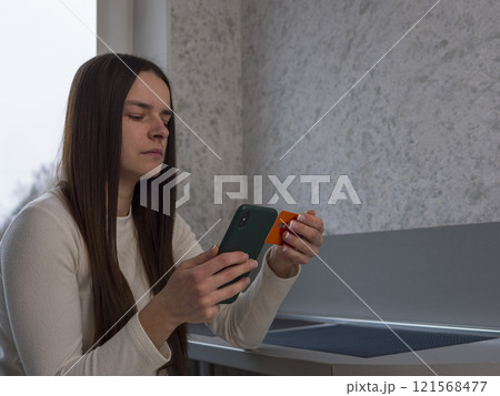 Young female completing mobile payment, gripping credit card and smartphone. Focused on digital transaction while seated near window, sunlight illuminating workspace Young female completing mobile payment, gripping credit card and smartphone. Focused on digital transaction while seated near window, sunlight illuminating workspace 121568477
