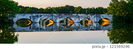 A stone inundation bridge from the late 18th century reflects in the calm waters of Vitek Pond in South Bohemia during a serene evening, surrounded by lush greenery. 121569296