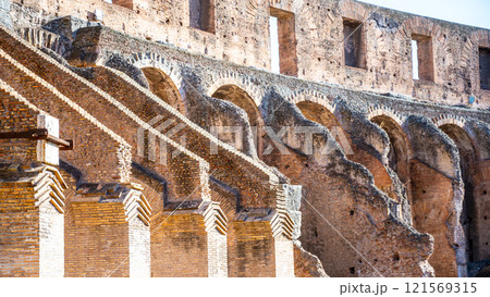 Visitors admire the intricate architecture of the Colosseum's interior in Rome. The ancient structure showcases impressive arches and historical remnants under the bright sky. 121569315