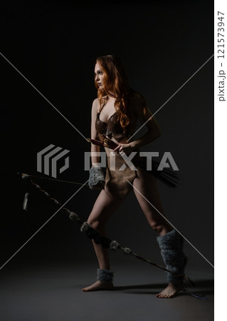 Woman in primitive attire posing with a bow and arrow in a dark studio setting 121573947