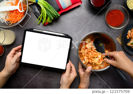 Man holding digital tablet while woman mixing kimchi in stainless steel bowl on a dark countertop 121575709
