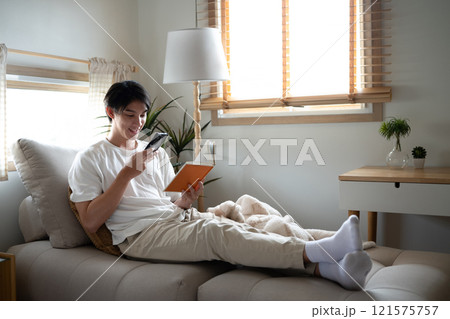 Carefree young adult man relaxing with a book and smartphone in cozy living room Carefree young adult man relaxing with a book and smartphone in cozy living room 121575757