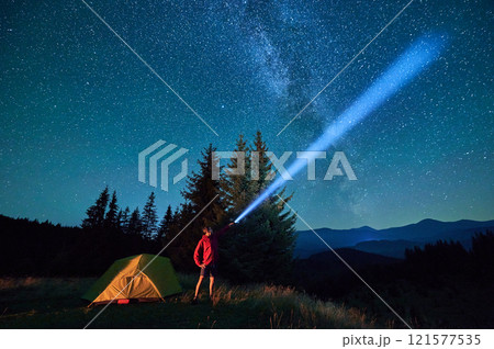 Man hiker stands under dazzling starry sky, shining flashlight into heavens. Milky Way stretches above, glowing tent nearby, creating magical and serene scene in peaceful mountain wilderness. Man hiker stands under dazzling starry sky, shining flashlight into heavens. Milky Way stretches above, glowing tent nearby, creating magical and serene scene in peaceful mountain wilderness. 121577535