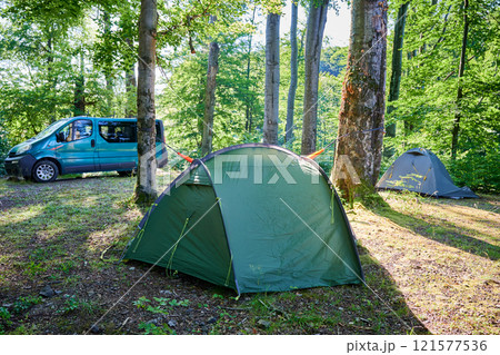 Multiple green tents in wooded campsite, with blue van parked nearby. Orange hammock strung between two trees. Sun filters through trees, casting dappled light across serene and inviting campsite. Multiple green tents in wooded campsite, with blue van parked nearby. Orange hammock strung between two trees. Sun filters through trees, casting dappled light across serene and inviting campsite. 121577536