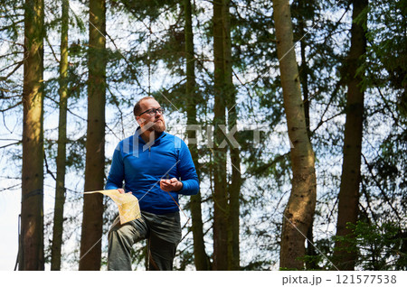 Man traveler in glasses stands in dense forest, holding map in one hand and compass in other. Tourist planning outdoor adventure or hiking route, amidst tall trees and natural surroundings. 121577538