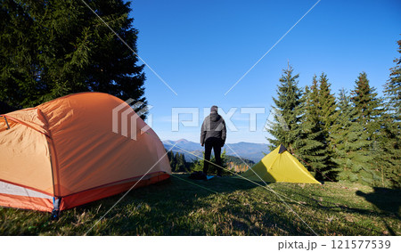 Back view of camper stands between orange and yellow tent, gazing at distant mountains under blue sky. Tall evergreen trees, casting shadows on grassy campsite. Morning light illuminates landscape. Back view of camper stands between orange and yellow tent, gazing at distant mountains under blue sky. Tall evergreen trees, casting shadows on grassy campsite. Morning light illuminates landscape. 121577539