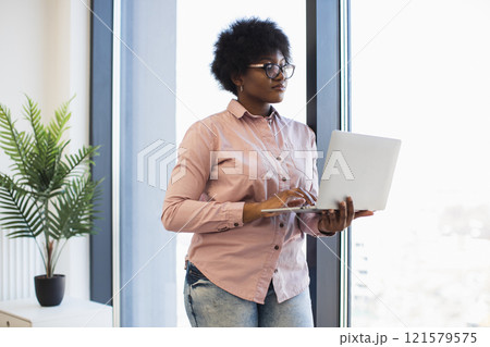 African American businesswoman stands by window using laptop. Young female in casual attire, focused on work. Modern office environment with natural light and plant decor. 121579575
