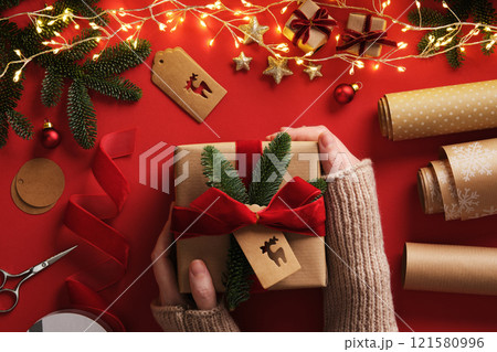 A woman's hands hold a wrapped gift. Table with handmade items, rolls of kraft brown paper, red velvet ribbon, labels, scissors. A woman's hands hold a wrapped gift. Table with handmade items, rolls of kraft brown paper, red velvet ribbon, labels, scissors. 121580996