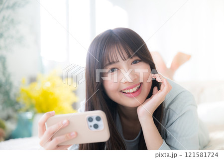 ベットで寝転がってスマホを持って笑顔のカメラ目線の若い女性のクローズアップ 春のイメージ ベットで寝転がってスマホを持って笑顔のカメラ目線の若い女性のクローズアップ 春のイメージ 121581724