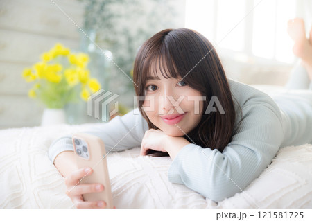 ベットで寝転がってスマホを持って笑顔のカメラ目線の若い女性のクローズアップ 春のイメージ ベットで寝転がってスマホを持って笑顔のカメラ目線の若い女性のクローズアップ 春のイメージ 121581725