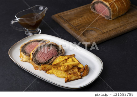 Beef Wellington with baked potato slice and demi-glace sauce. Traditional festive dish of beef tenderloin with mushrooms and Parma ham baked in puff pastry. Selective focus, close-up. 121582789