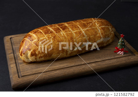 Whole beef Wellington on a wooden board. A small toy Christmas tree nearby. Traditional festive dish of beef tenderloin with mushrooms and Parma ham baked in puff pastry. Selective focus, close-up. Whole beef Wellington on a wooden board. A small toy Christmas tree nearby. Traditional festive dish of beef tenderloin with mushrooms and Parma ham baked in puff pastry. Selective focus, close-up. 121582792