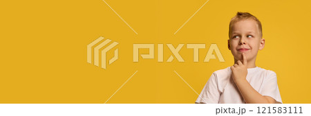 Boy strikes thoughtful pose, hand on chin, smiling slyly, dressed in white shirt, exuding curiosity against bright yellow background. 121583111