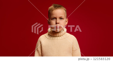 Boy frowns slightly, creating pouting expression, wearing knitted sweater, standing against deep red background. 121583120