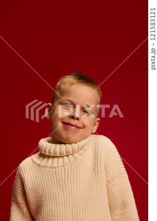 Boy smiles warmly, wearing knitted sweater, tilting head slightly, expressing confidence against deep red background. 121583121