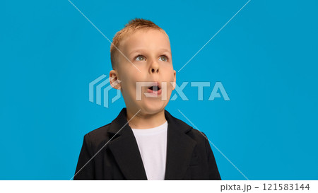 Surprised boy in black blazer and white shirt, wide-eyed, making an exaggerated expression of amazement, radiating curiosity against bright blue background. 121583144