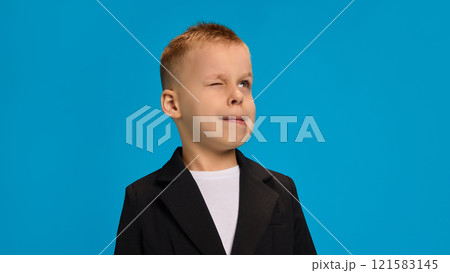 Playful boy in black blazer and white shirt, winking with lips pursed slightly, showcasing a cheeky, fun attitude against bright blue background. Playful boy in black blazer and white shirt, winking with lips pursed slightly, showcasing a cheeky, fun attitude against bright blue background. 121583145