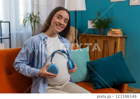 Pregnant woman sitting on home sofa listening music putting headphones on her tummy with child baby Pregnant woman sitting on home sofa listening music putting headphones on her tummy with child baby 121586149