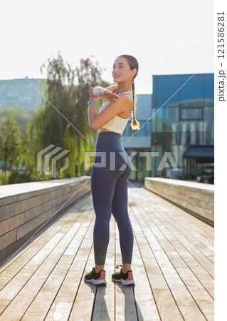 Smiling young sportswoman stretching hands shoulders and muscles workout in city park on sunny day Smiling young sportswoman stretching hands shoulders and muscles workout in city park on sunny day 121586281