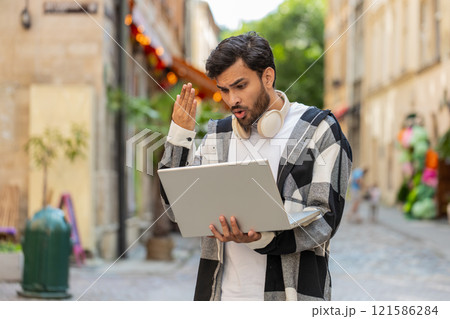 Shocked Indian man using laptop becoming surprised sudden lottery results bad news in city street Shocked Indian man using laptop becoming surprised sudden lottery results bad news in city street 121586284