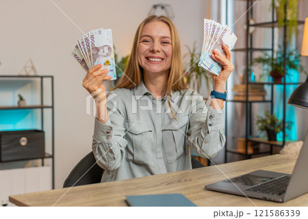 Excited businesswoman freelancer holding fan of Ukrainian hryvnias UAH cash dancing at home office 121586339