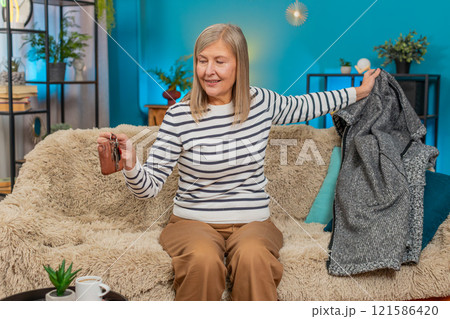 Mature woman holding apartment keys, joyfully entering home and sitting on sofa, removing jacket 121586420