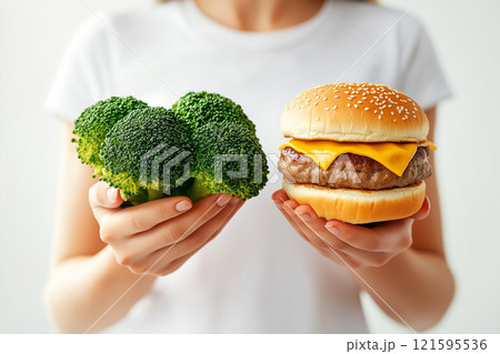 Woman holding broccoli and a burger. A choice between healthy nutrition and junk food, diet, nutrition, healthy lifestyle concept. Dieting, food choices, cheat meal. 121595536