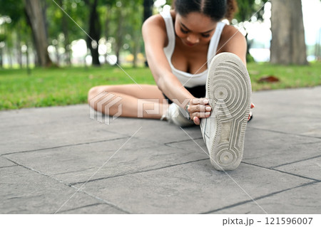 Young sportswoman stretching legs on a pathway preparing for run or workout in the park 121596007