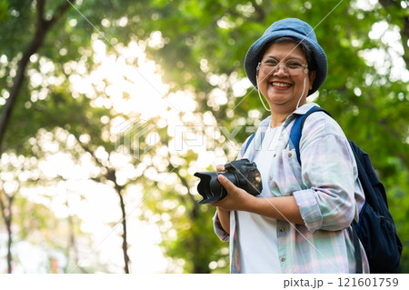 Portrait of Asian mature woman holding a camera and backpack behind her back, an Asia active senior woman enjoying nature in park. Standing on a trail in a forest outdoors. Enjoying active travel trip 121601759