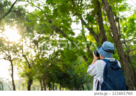 Portrait of Asian mature woman holding a camera and backpack behind her back, an Asia active senior woman enjoying nature in park. Standing on a trail in a forest outdoors. Enjoying active travel trip 121601760