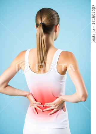 Woman experiencing lower back pain while standing against a light blue background during daylight hours Woman experiencing lower back pain while standing against a light blue background during daylight hours 121602587