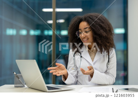 A female doctor sitting in her office and engaging in a video consultation through her laptop. She exhibits a professional demeanor and uses virtual technology for communication with patients. A female doctor sitting in her office and engaging in a video consultation through her laptop. She exhibits a professional demeanor and uses virtual technology for communication with patients. 121603955