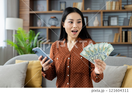 Excited woman holds cash while checking her phone, indicating online earnings success from home. Asian woman sits on cozy sofa in modern living room, expressing financial achievement and happiness. Excited woman holds cash while checking her phone, indicating online earnings success from home. Asian woman sits on cozy sofa in modern living room, expressing financial achievement and happiness. 121604311