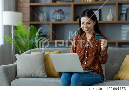 Asian woman shopping online using laptop and credit card, sitting comfortably on sofa in home setting. Concept of convenience, technology, and e-commerce in everyday life. Asian woman shopping online using laptop and credit card, sitting comfortably on sofa in home setting. Concept of convenience, technology, and e-commerce in everyday life. 121604378